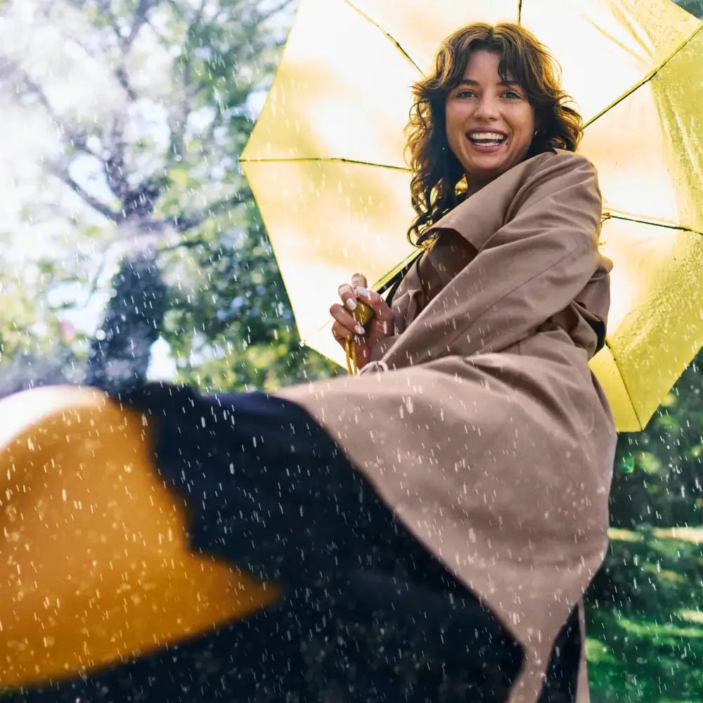Lächelnde Person, die den gelben Regenschirm „NATURAL ELEMENTS Immun & Feelgood Bundle“ im Regen hält, trägt einen braunen Mantel und vor einem unscharfen Hintergrund mit grünen Bäumen.